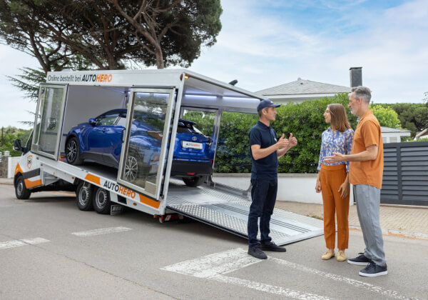 Autohero delivery driver talking to a customer couple, in the back a Autohero glass-truck with lowered back ready to unload a car.